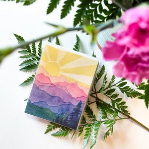 May include: A square greeting card featuring a watercolour illustration of a mountain landscape at sunrise. The scene displays layered purple and pink mountain peaks beneath a bright yellow sun with radiating beams, and dark green pine trees at the base.