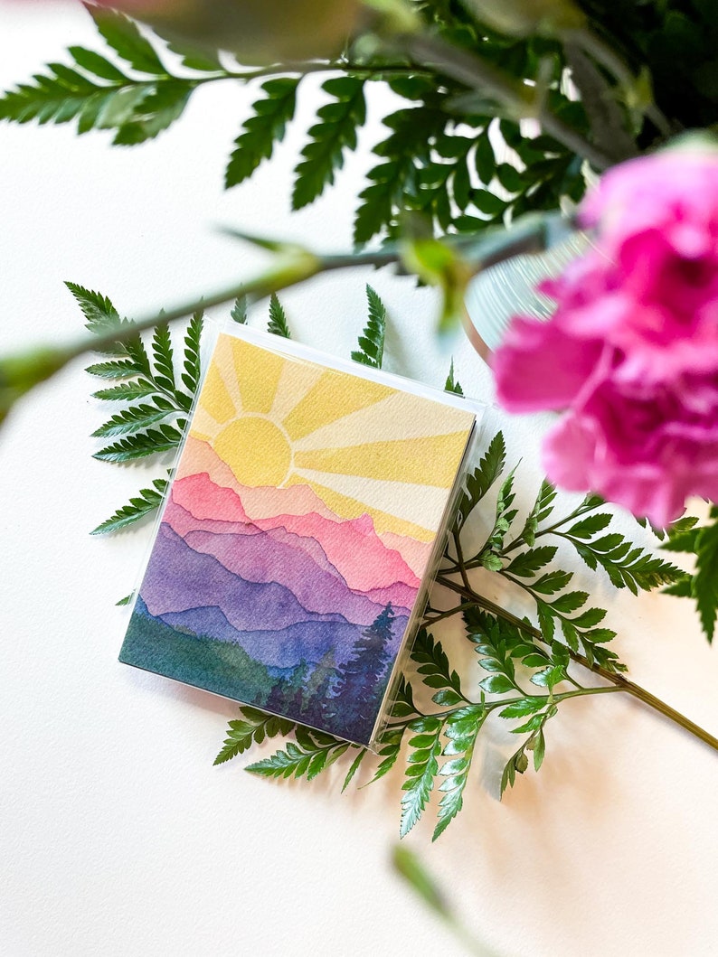 May include: A square greeting card featuring a watercolour illustration of a mountain landscape at sunrise. The scene displays layered purple and pink mountain peaks beneath a bright yellow sun with radiating beams, and dark green pine trees at the base.