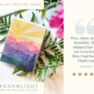 May include: A square greeting card featuring a watercolour-style illustration of a sunset over layered mountain ranges in shades of yellow, pink, purple, and blue, with dark green pine trees in the foreground. The text reads SUN SET GREETING CARDS.