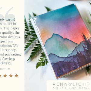 May include: A set of greeting cards in a clear plastic sleeve featuring a watercolour landscape of mountains and pine trees at sunset with shades of orange, pink, purple, and teal. The text on the left reads: Lovely cards! Even better in person.