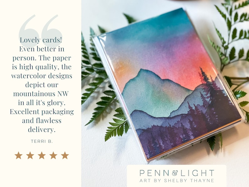 May include: A set of greeting cards in a clear plastic sleeve featuring a watercolour landscape of mountains and pine trees at sunset with shades of orange, pink, purple, and teal. The text on the left reads: Lovely cards! Even better in person.