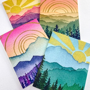 May include: Four rectangular greeting cards featuring watercolour-style landscape illustrations. Each card depicts a sun rising or setting over layered mountains and evergreen forests, accented with gold-coloured lines in the sky and sun rays.