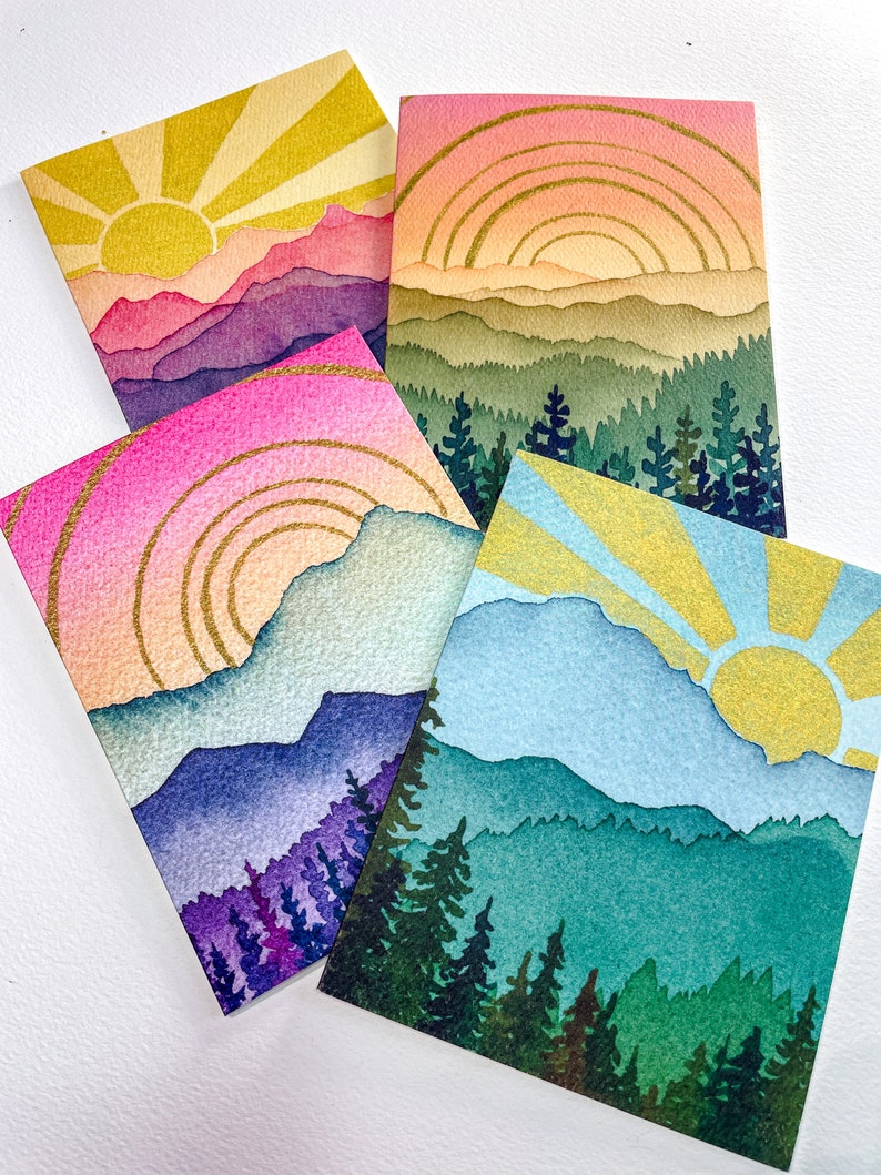 May include: Four rectangular greeting cards featuring watercolour-style landscape illustrations. Each card depicts a sun rising or setting over layered mountains and evergreen forests, accented with gold-coloured lines in the sky and sun rays.