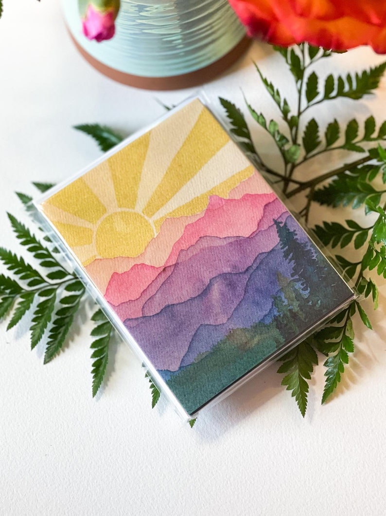 May include: A rectangular deck of cards encased in a clear plastic box featuring a watercolour illustration of a sunrise over layered mountain ranges in shades of yellow, pink, purple, and blue, with dark green pine trees on the right side.