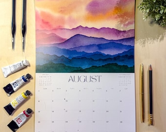 May include: A wall calendar for August 2027 featuring a watercolour mountain landscape with a sunset sky in shades of orange, pink, and purple. The calendar includes a grid for dates, the text AUGUST 2027, and the artist name Shelby Thayne Art.