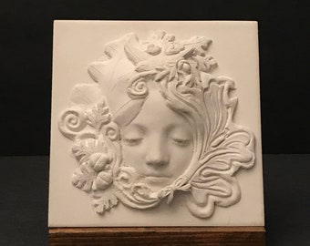 May include: A white relief sculpture of a serene face surrounded by foliage, including leaves and acorns. The sculpture is mounted on a small, rectangular wooden base. The face has closed eyes and a gentle expression.