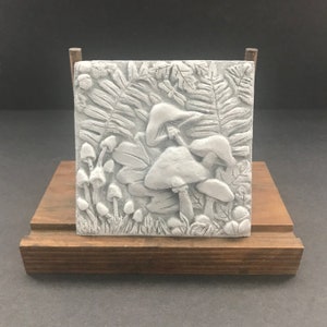 May include: A square, gray relief sculpture of mushrooms and foliage, including ferns and leaves. The artwork is mounted on a wooden stand. The sculpture features detailed textures and a naturalistic design, evoking a forest scene.