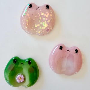 Peut inclure: Trois ornements en résine en forme de grenouille avec des surfaces lisses et brillantes. L'un est rose clair avec des paillettes irisées, l'un est rose clair uni et l'un est vert avec une petite fleur rose. Chacun a deux yeux noirs et une petite bouche.