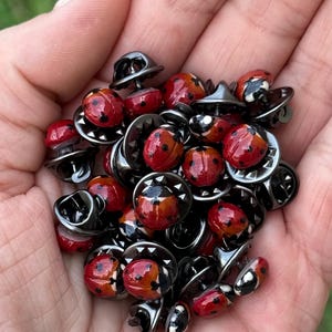 Peut inclure: Une collection de petites broches en forme de coccinelle avec des carapaces rouges arrondies et des points noirs distincts. Chaque broche présente une finition brillante et un support métallique sombre avec un fermoir sécurisé.