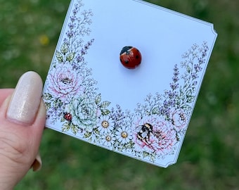 May include: A small, round, red ladybug pin with black spots attached to a square white display card. The card features a decorative floral border illustration with pink peonies, white daisies, lavender sprigs, and a small bumblebee graphic.