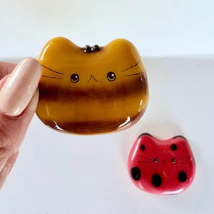 Peut inclure: Deux objets décoratifs en forme de chat. L'un est de couleur ambre avec des moustaches et des yeux noirs. L'autre est rouge avec des taches et des moustaches noires. Les deux ont une finition brillante et sont sur fond blanc.