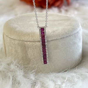 Puede incluir: Un collar de plata con un colgante que presenta una fila vertical de piedras preciosas rojas cuadradas rodeadas de piedras preciosas blancas más pequeñas.