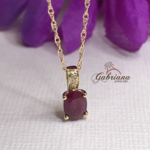 Könnte beinhalten: Eine goldene Kettenkette mit einem Anhänger mit einem roten ovalen Rubin-Edelstein, der von kleinen Diamanten umgeben ist.