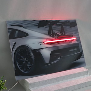 Puede incluir: Un Porsche 911 GT3 RS plateado con una luz trasera roja y un alerón trasero negro. El coche tiene un difusor trasero negro y el texto "PORSCHE" y "DRIVEN TO FORCE" en la parte trasera.