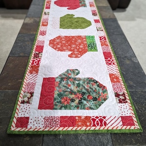 以下が含まれることがあります： 緑と赤の縁取りが付いた白いキルトのテーブルランナー。ランナーには、クリスマスをテーマにした異なる生地のパターンで、ミトン型のアップリケが4つ付いています。