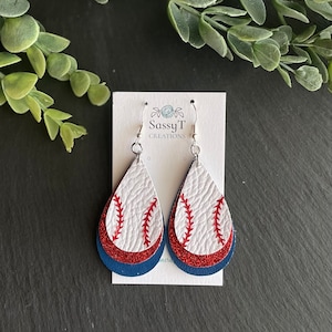 May include: A pair of dangle earrings with a layered design. The top layer is white leather with a red baseball stitch design. The middle layer is red glitter and the bottom layer is navy blue leather. The earrings are attached to silver hooks. The card behind the earrings says "Sassy T Creations".