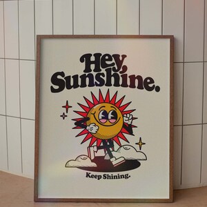 May include: A framed print with a cartoon sun character with the text "Hey, Sunshine. Keep Shining." in black and white lettering on a white background.