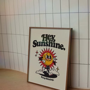 May include: A framed print with a yellow cartoon sun with a smiling face and red rays. The sun is walking on clouds and the text "Hey Sunshine. Keep Shining." is below the sun.