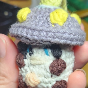May include: A crocheted mushroom-shaped plushie with a white body, brown accents, and a purple hat with yellow accents.