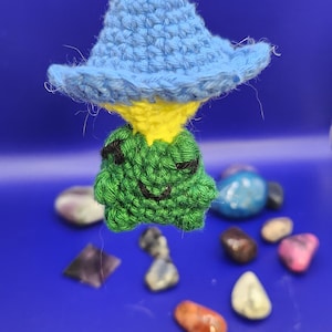 Puede incluir: Una criatura verde de crochet con una cara amarilla que lleva un sombrero azul y amarillo. La criatura está colgada de una cuerda amarilla y está rodeada de piedras de colores.