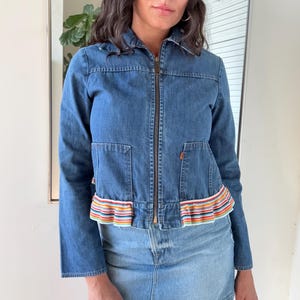 May include: A blue denim jacket with a zip-up front, two front pockets, and long sleeves. The jacket features a colorful striped ruffle detail at the waist. The model is also wearing a denim skirt.