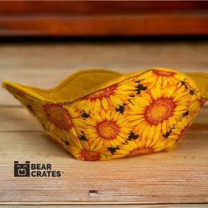 May include: A yellow bowl cozy with a sunflower pattern. The cozy is made of fabric and has a square shape with curved edges. The interior is a solid yellow color. The logo "Bear Crate" is visible.