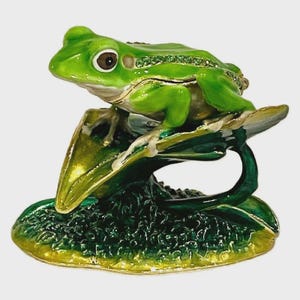 May include: A decorative frog figurine in shades of green, perched on a lily pad. The frog has brown eyes and is embellished with small, sparkling accents. The lily pad is detailed with green and yellow hues, creating a nature-inspired trinket.