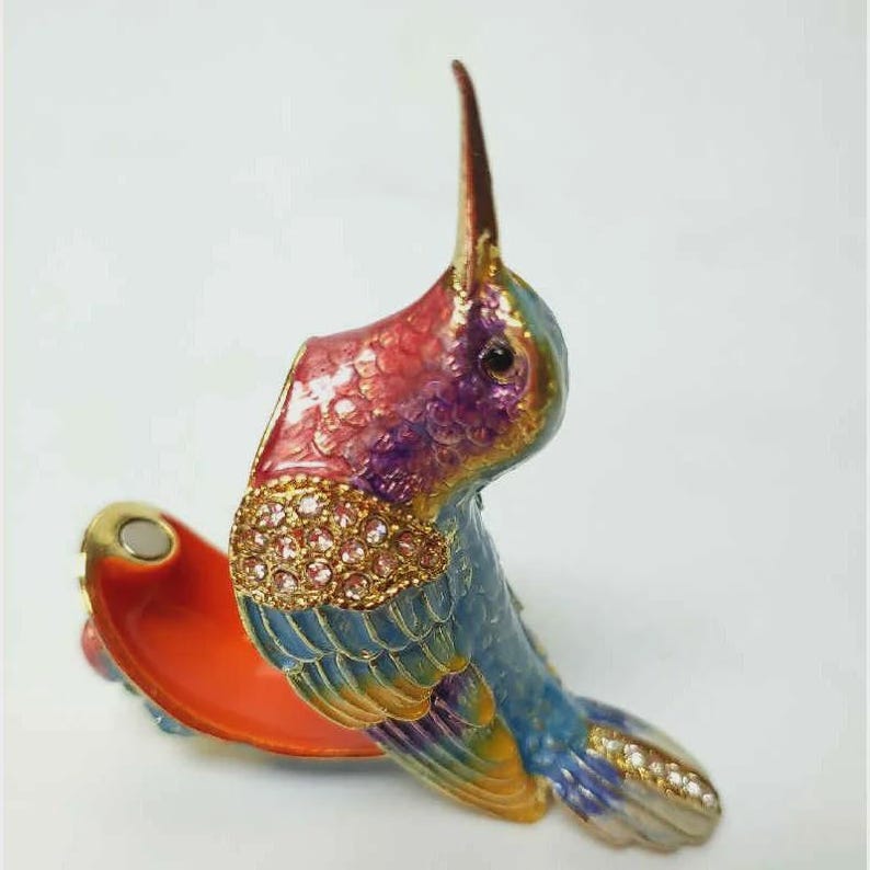 May include: A decorative hummingbird trinket box. The box is open, revealing an orange interior. The hummingbird is painted in shades of blue, purple, red, and gold, with a gold band of pink rhinestones around its neck. The bird's beak is long and dark.