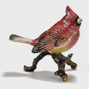 May include: A red cardinal bird figurine perched on a textured dark brown branch. The bird features black wing accents, a gold-toned beak, and a row of small clear crystals along the side of its body. The surface has a glossy, enameled finish.