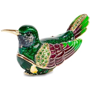May include: A hummingbird figurine crafted with metallic green enamel, featuring a long gold-toned beak, wings accented with deep magenta and green enamel, and a patch of small, sparkling green crystals on the upper wing section.