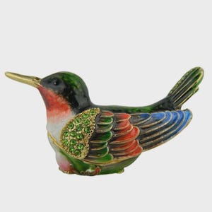 May include: A decorative hummingbird figurine with a gold beak and accents. The bird features a green head, black back, red and blue wings, and a gold-colored chest adorned with green gemstones. The figurine is a small, detailed piece.