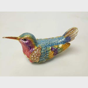 May include: A colourful hummingbird ornament with a long beak. The bird has a blue head and back, a pink chest, and yellow and purple wings. The wings are detailed with gold and rhinestones, creating a decorative piece.