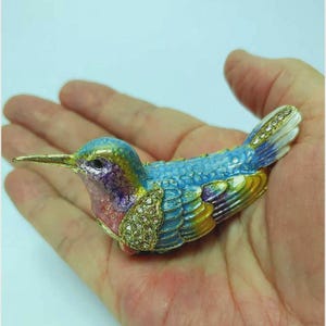May include: A decorative hummingbird ornament, featuring a gold beak and jewel-encrusted chest. The bird's body is painted in shades of blue, green, yellow, and purple. The wings and tail are detailed with gold and crystal-like accents.