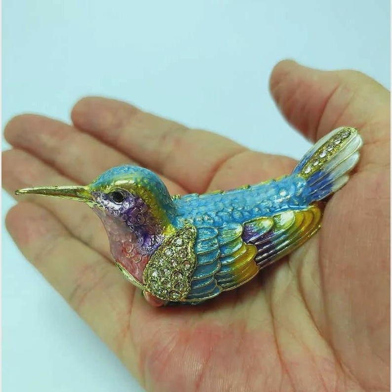 May include: A decorative hummingbird ornament, featuring a gold beak and jewel-encrusted chest. The bird's body is painted in shades of blue, green, yellow, and purple. The wings and tail are detailed with gold and crystal-like accents.