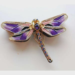 May include: A decorative dragonfly brooch with gold-tone metalwork. The wings are painted in shades of purple, pink, and white, accented with small crystals. The body features a large purple gemstone and smaller blue and pink gems.