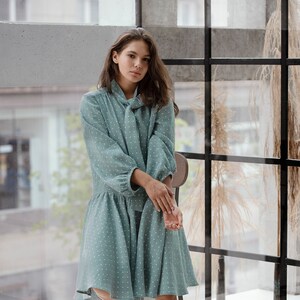 May include: A woman wearing a teal blue dress with white polka dots. The dress has a tie neck and long sleeves. She is sitting on a wooden stool in front of a window.