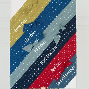 May include: A wooden easel displaying fabric swatches with white polka dots in various colours, including green, blue, mint, navy blue, red, and denim blue. Each swatch has a label with the colour name and the words "Dots" written in white.