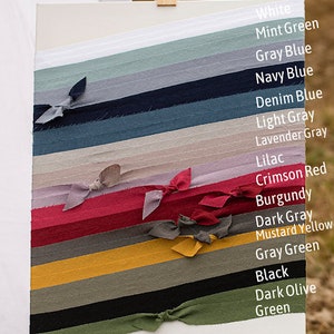 May include: A white canvas with 16 fabric swatches tied in knots. The swatches are arranged vertically from top to bottom in the following colours: white, mint green, grey blue, navy blue, denim blue, light grey, lavender grey, lilac, crimson red, burgundy, dark grey, mustard yellow, grey green, black, dark olive green, and green.