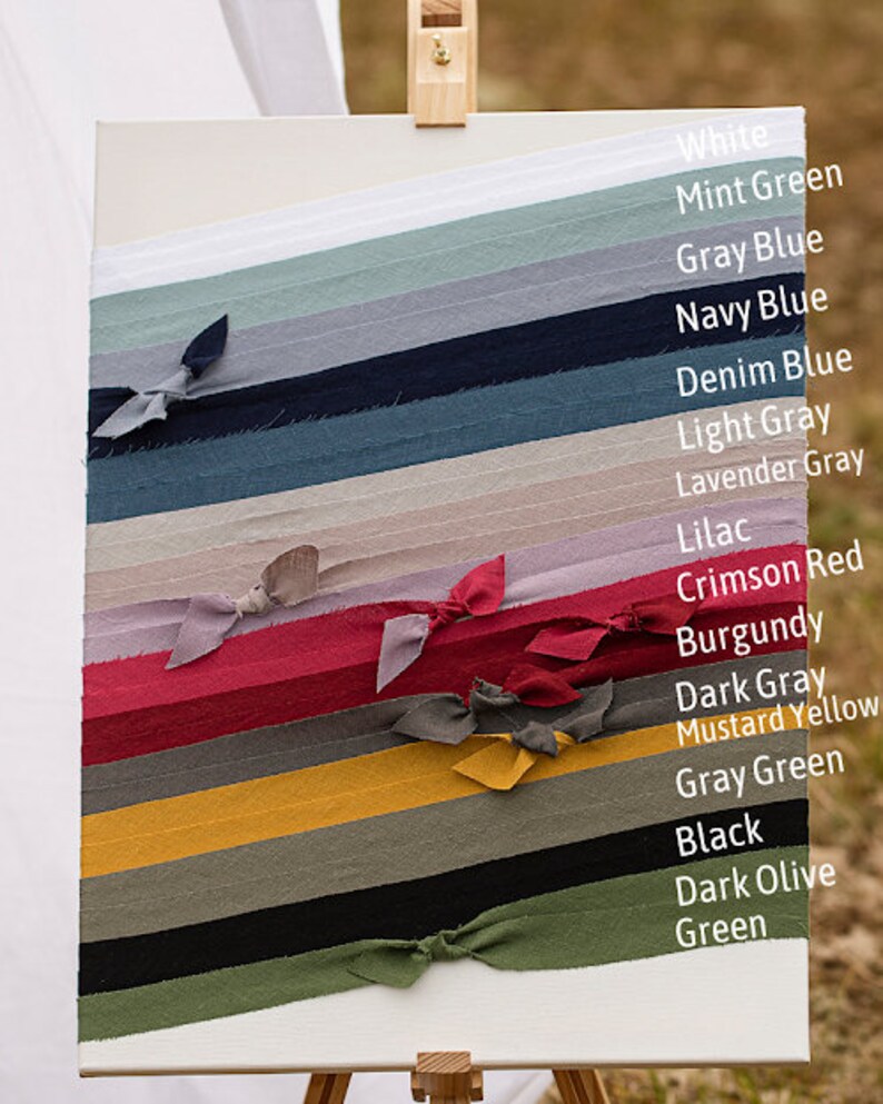 May include: A white canvas with 16 fabric swatches tied in knots. The swatches are arranged vertically from top to bottom in the following colours: white, mint green, grey blue, navy blue, denim blue, light grey, lavender grey, lilac, crimson red, burgundy, dark grey, mustard yellow, grey green, black, dark olive green, and green.