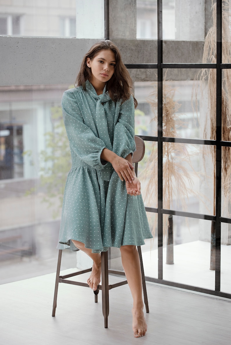 May include: A woman wearing a teal blue dress with white polka dots. The dress has a tie neck and long sleeves. She is sitting on a wooden stool in front of a window.