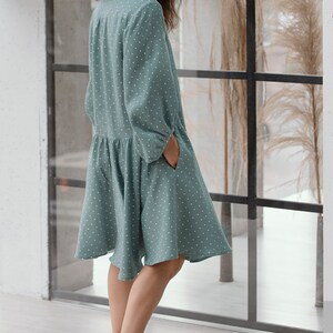 May include: A woman wearing a teal blue dress with white polka dots. The dress has a collared neckline and long sleeves. The woman is standing in front of a window with a view of a grassy field.