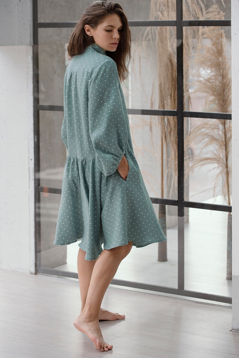 May include: A woman wearing a teal blue dress with white polka dots. The dress has a collared neckline and long sleeves. The woman is standing in front of a window with a view of a grassy field.