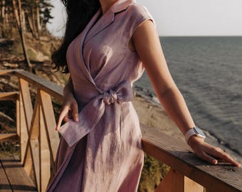 May include: A light purple wrap dress with a bow at the waist. The dress has short sleeves and a collared neckline. The person in the image is wearing sunglasses and a watch. The background includes a wooden railing and a view of the ocean.