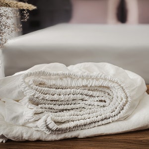 Puede incluir: Una sábana bajera blanca está enrollada y doblada sobre una superficie de madera. La sábana está hecha de una tela suave y natural y tiene una textura lisa y uniforme.