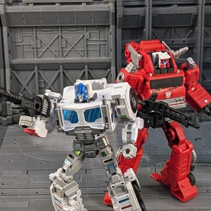 May include: Two toy robots, one white and one red, stand in front of a gray background. The white robot has blue accents and is holding a black gun. The red robot is holding a black gun as well.
