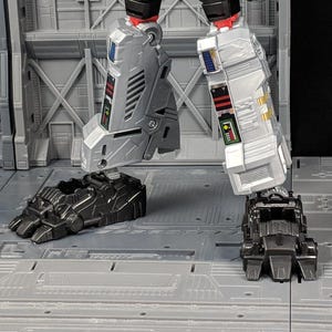 May include: Close-up of a robot's feet. The feet are black and have a detailed design. The robot's legs are silver and have a complex design with black and red accents.
