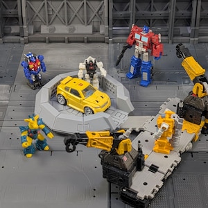 May include: A collection of toy robots and a yellow car are arranged on a gray platform. The robots are in various colors, including red, blue, yellow, and white. One robot is holding a black weapon. The scene is set against a gray background.