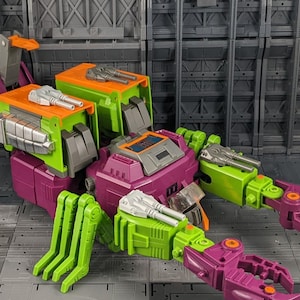 May include: A multi-colored toy robot with a purple body, green and orange accents, and silver weaponry. The robot has articulated limbs and a detailed design, suggesting a collectible or action figure.
