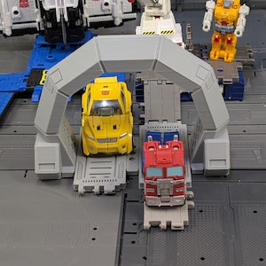 May include: A yellow car and a red truck toy are positioned under a gray archway. The toys are on a gray platform with other toy components in the background. Other toy figures are visible.
