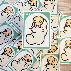 Puede incluir: Una pila de pegatinas con un personaje de dibujos animados con una cabeza amarilla y un cuerpo blanco. El personaje está enroscado en una bola y tiene una expresión linda en su rostro. Las pegatinas están impresas en cartulina blanca con un borde verde.